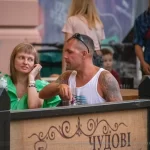 Фотография мужчины с татуировкой на руке, сидящего за столиком в уличном кафе с женщиной. На столе напитки и закуски.