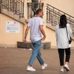 Фотография мужчины в светлой одежде с татуировкой на руке, идущего мимо памятника на городской улице.