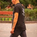 Фотография мужчины в кепке и темной одежде с татуировкой волка на руке, идущего по улице.