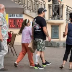 Фотография мужчины с татуировкой на руке, стоящего на улице. На заднем плане виден памятник и прохожие.