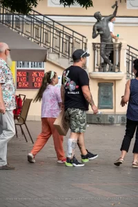 Фотография мужчины с татуировкой на руке, стоящего на улице. На заднем плане виден памятник и прохожие.
