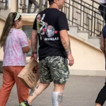 Фотография мужчины с татуировкой на руке, идущего по улице с пакетом покупок. Татуировка заметна на предплечье, а на лице мужчины выражение задумчивости.