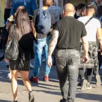 Фотография мужчины с полным рукавом татуировок, идущего по улице в окружении людей.