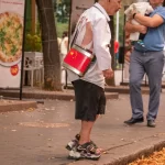 Фотография пожилого уличного музыканта с татуировкой на стопе, играющего на барабане с китайской символикой на фоне городской улицы.