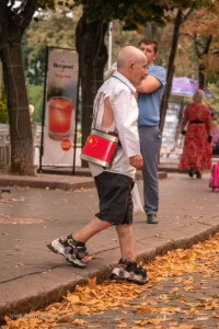 Фотография пожилого уличного музыканта с татуировкой на стопе, играющего на барабане с китайской символикой на фоне городской улицы.