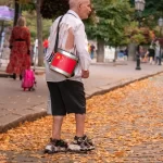 Фотография уличного музыканта с красным барабаном и рваной рубашкой, идущего по осенней аллее. Необычный пример использования тела как холста для тату.