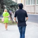 Фотография мужчины на электросамокате с татуировкой на руке и девушки, идущей рядом. Городской пейзаж и динамичная сцена.