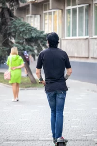 Фотография мужчины на электросамокате с татуировкой на руке и девушки, идущей рядом. Городской пейзаж и динамичная сцена.