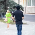 Фотография мужчины на электросамокате с татуировкой на руке и девушки, идущей рядом. Городской пейзаж и динамичная сцена.