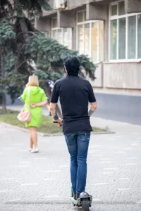 Фотография мужчины на электросамокате с татуировкой на руке и девушки, идущей рядом. Городской пейзаж и динамичная сцена.