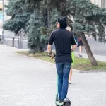 Фотография мужчины, едущего на электросамокате, с видимой татуировкой на руке. Городской пейзаж и динамичная поза.