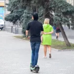 Фотография мужчины на электросамокате с татуировкой на руке и девушки, идущей рядом. Городской пейзаж и динамичная сцена.