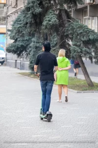 Фотография мужчины на электросамокате с татуировкой на руке и девушки, идущей рядом. Городской пейзаж и динамичная сцена.