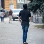 Фотография мужчины, едущего на электросамокате, с видимой татуировкой на руке. Городской пейзаж и динамичная поза.