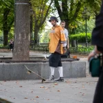 Фотография скейтбордиста с татуировками на руках, выполняющего трюк в парке. Он одет в оранжевую футболку и черные шорты.