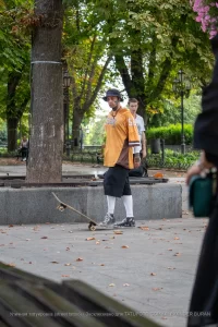 Фотография скейтбордиста с татуировками на руках, выполняющего трюк в парке. Он одет в оранжевую футболку и черные шорты.