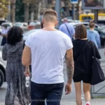 Фотография мужчины с татуировкой на руке, идущего по улице в белой футболке и джинсах, спиной к камере.