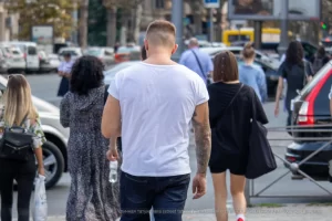 Фотография мужчины с татуировкой на руке, идущего по улице в белой футболке и джинсах, спиной к камере.