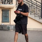 Фотография мужчины с татуировкой на икре, идущего по улице. Татуировка выполнена в ярком стиле с использованием красного и черного цветов. Отличный пример уличной татуировки.