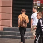 Фото мужчины с татуировками на руках, идущего по улице с рюкзаком. Видны фрагменты рисунков с цветами и абстрактными элементами.