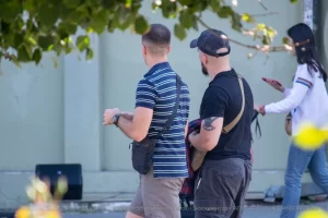 Фотография мужчины с татуировкой на руке, идущего по улице. На заднем плане видны деревья и люди.