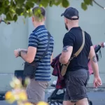 Фотография мужчины с татуировкой на руке, идущего по улице. На заднем плане видны деревья и люди.
