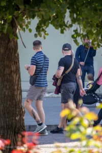 Фотография мужчины с татуировкой на руке, идущего по улице с двумя другими людьми. На заднем плане видны деревья и цветы.