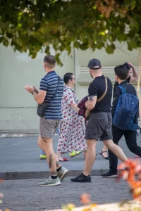 Фотография мужчины с татуировкой на руке, идущего по улице с двумя другими людьми. На заднем плане видны деревья и цветы.