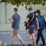 Фотография мужчины с татуировкой на ноге, идущего по улице с двумя другими людьми. На заднем плане видны деревья и цветы.