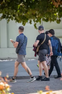 Фотография мужчины с татуировкой на ноге, идущего по улице с двумя другими людьми. На заднем плане видны деревья и цветы.