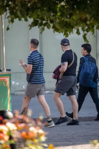Фотография мужчины с татуировкой на руке, идущего по улице с двумя другими людьми. На заднем плане видны деревья и цветы.