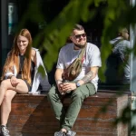 Фотография мужчины с татуировками на руке и рыжеволосая девушка, сидящих на деревянном ящике. Мужчина одет в белую футболку и зеленые брюки.