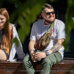 Фотография мужчины с татуировками на руке и рыжеволосая девушка, сидящих на деревянном ящике. Мужчина одет в белую футболку и зеленые брюки.