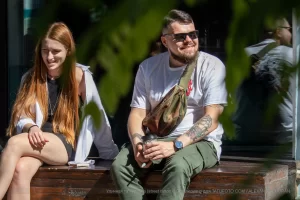 Фотография мужчины с татуировками на руке и рыжеволосая девушка, сидящих на деревянном ящике. Мужчина одет в белую футболку и зеленые брюки.