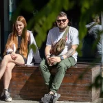 Фотография мужчины с татуировками на руке и рыжеволосая девушка, сидящих на деревянном ящике. Мужчина одет в белую футболку и зеленые брюки.