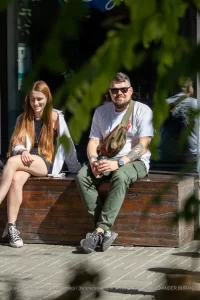 Фотография мужчины с татуировками на руке и рыжеволосая девушка, сидящих на деревянном ящике. Мужчина одет в белую футболку и зеленые брюки.