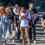 Фотография мужчины с татуировками на руках, идущего по улице и смотрящего в смартфон. Он одет в белую футболку и светлые брюки.