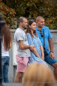 Фото мужчины с татуировкой на руке, стоящего рядом с женщиной и другим мужчиной. Татуировка представляет собой спираль.