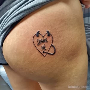 Фото тату: небольшая татуировка с надписью "Spank Me" в форме сердца с рогами на бедре девушки.