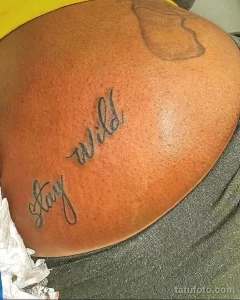 Фото татуировки на плече с надписью "Stay Wild" выполненной тонким шрифтом. Рисунок выполнен в черном цвете.
