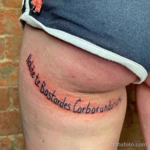 Фото тату: фраза на латыни 'Nolite te Bastardes Carborundorum' на женском бедре. Стильная и оригинальная надпись татуировки.