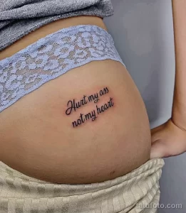 Фото тату надписи 'Hurt my ass not my heart' на бедрах. Черный контур, минималистичный стиль, женственная татуировка.