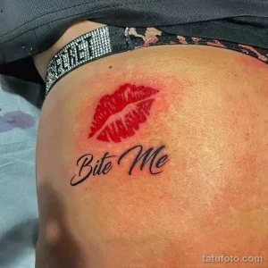 Фото тату на плече: реалистичное изображение красных губ с надписью 'Bite Me' выполненное черными чернилами.