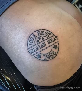 Фото тату: стилизованная печать '100% BEST BELGIAN MEAT 1988' на спине мужчины, выполненная в технике blackwork.