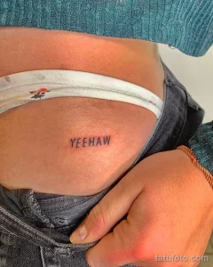 Фото тату надписи 'Yeehaw' на боку. Простая, минималистичная татуировка черным цветом, выполненная тонкой линией.