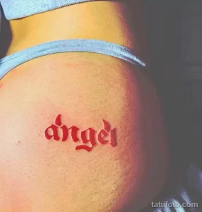 Фото минималистичной татуировки надписи 'angel' на плече, выполненной красным цветом в готическом стиле.