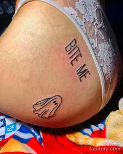 Фото тату с изображением маленького привидения и надписью 'BITE ME' на бедре. Рисунок выполнен в черном цвете в минималистичном стиле.