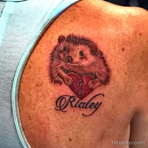 Фото тату: милый ёжик, держащий разбитое сердце с именем 'Rialey' на плече. Цветная татуировка с использованием нежных оттенков.