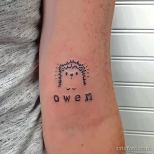 Фото тату маленького ежика с надписью 'Owen' под ним, на предплечье. Простой и минималистичный рисунок в черном цвете.