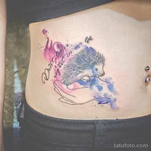 Фото акварельной татуировки ежика с надписью "Don't leave regret in our life" на боку. Рисунок выполнен в мягких тонах с использованием техники акварели.